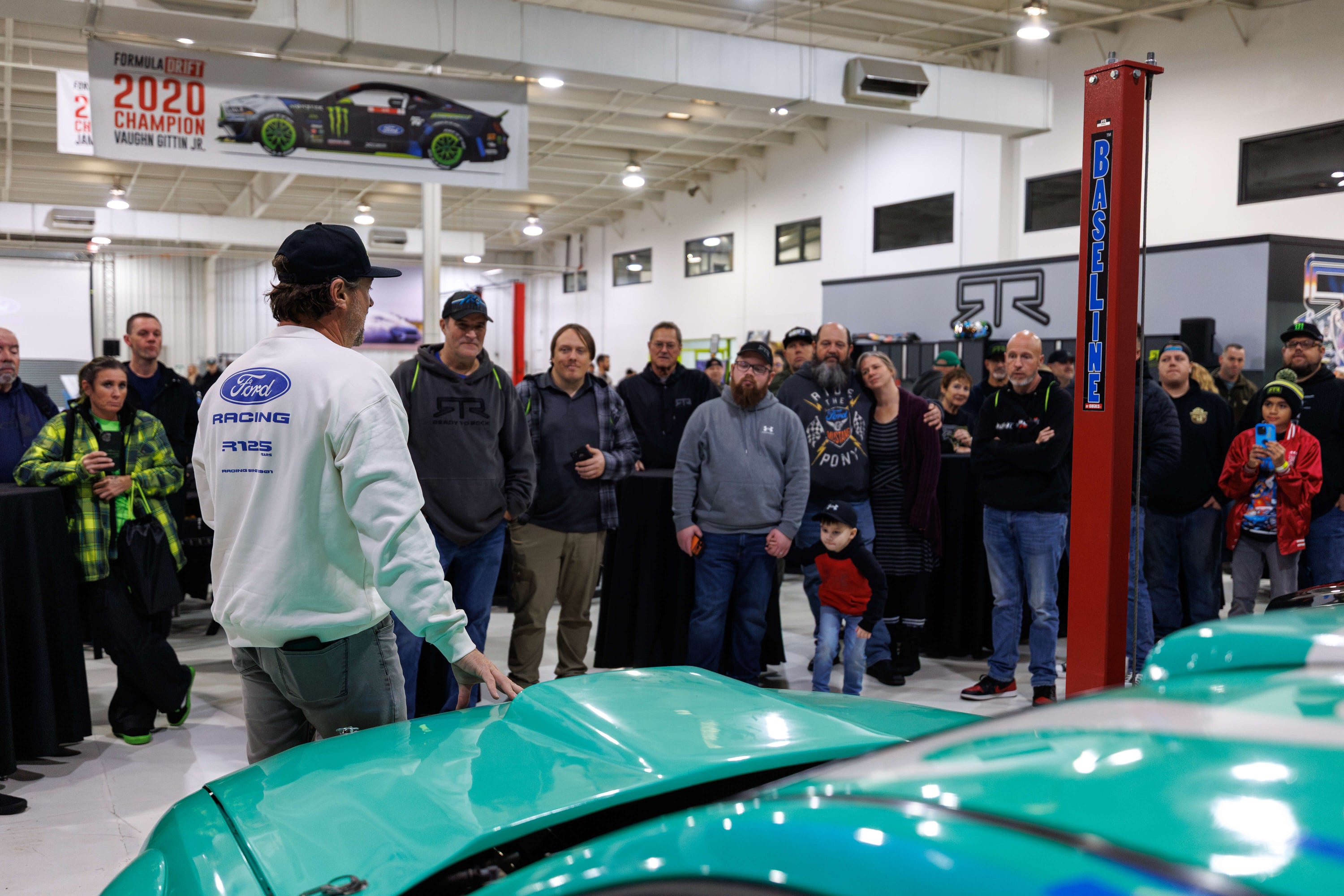 Group of people gathered around a teal car in a showroom listening to Vaughn Gittin Jr.