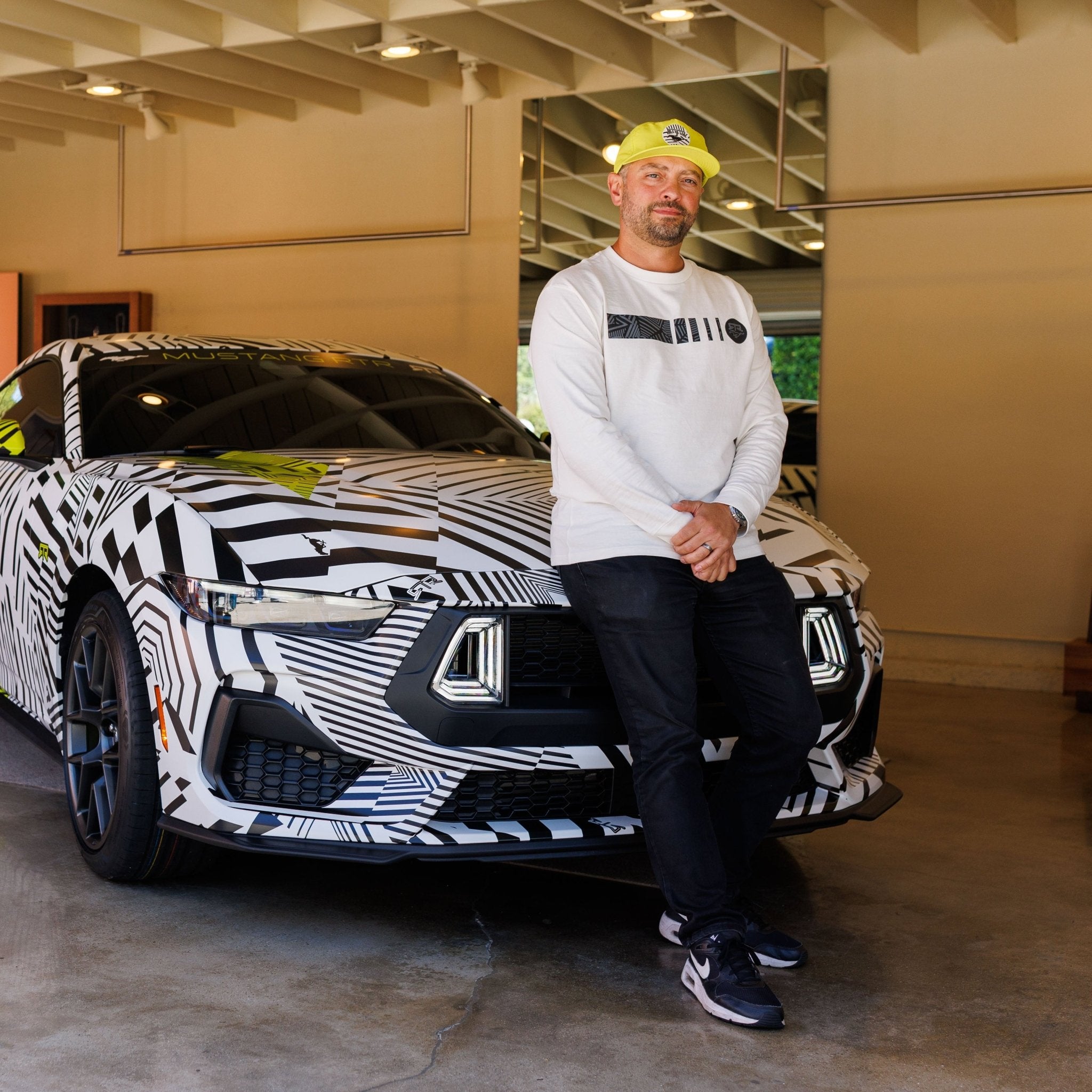 Man wearing the Ford Mustang x RTR Collection 5 Panel Unstructured Snapback Hat, leaning on a camouflaged sports car in a showroom.