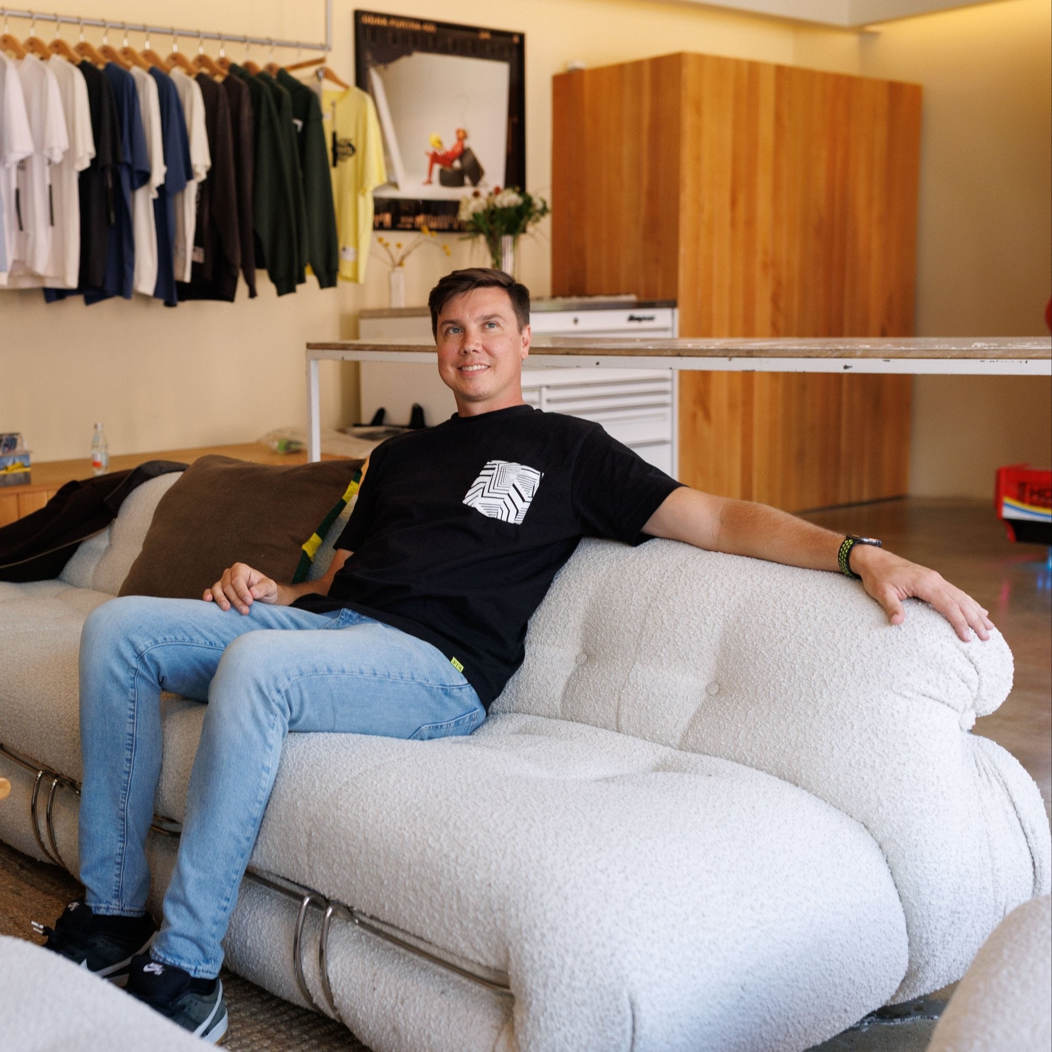 Man lounges on a white boucle sofa wearing the Ford Mustang x RTR Collection Pocket T-Shirt, featuring RTR's dazzle camo design pocket in a minimalist showroom.