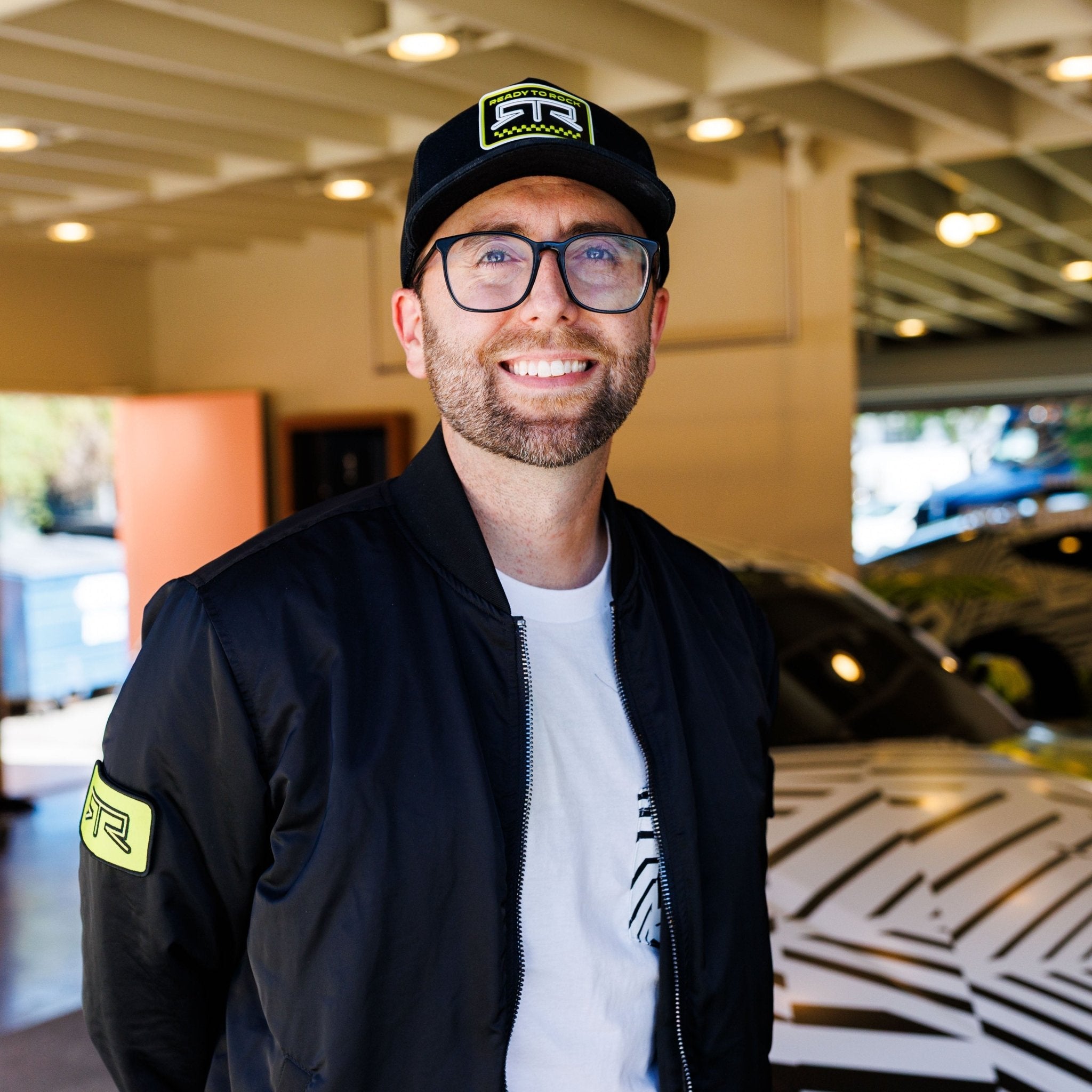 Man wearing Ford Mustang x RTR Collection Stealth Jacket, featuring a sleek black design, showcases modern utility and style amidst a garage setting with a camouflaged car.