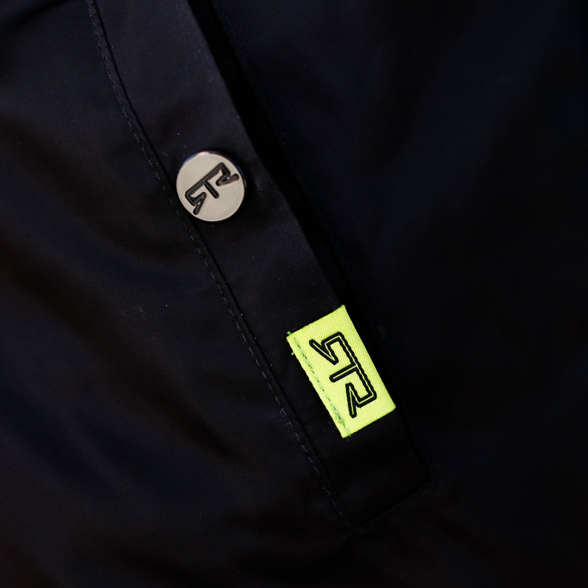 Close-up of Ford Mustang x RTR Collection Stealth Jacket, showing a metal snap button with “TR” logo and neon-yellow tag, reflecting performance-inspired design.