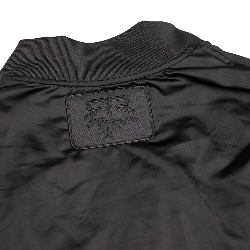 Black bomber jacket's upper back with ribbed collar, featuring a rectangular patch with RTR and horse logo. Ford Mustang x RTR Collection Stealth Jacket.