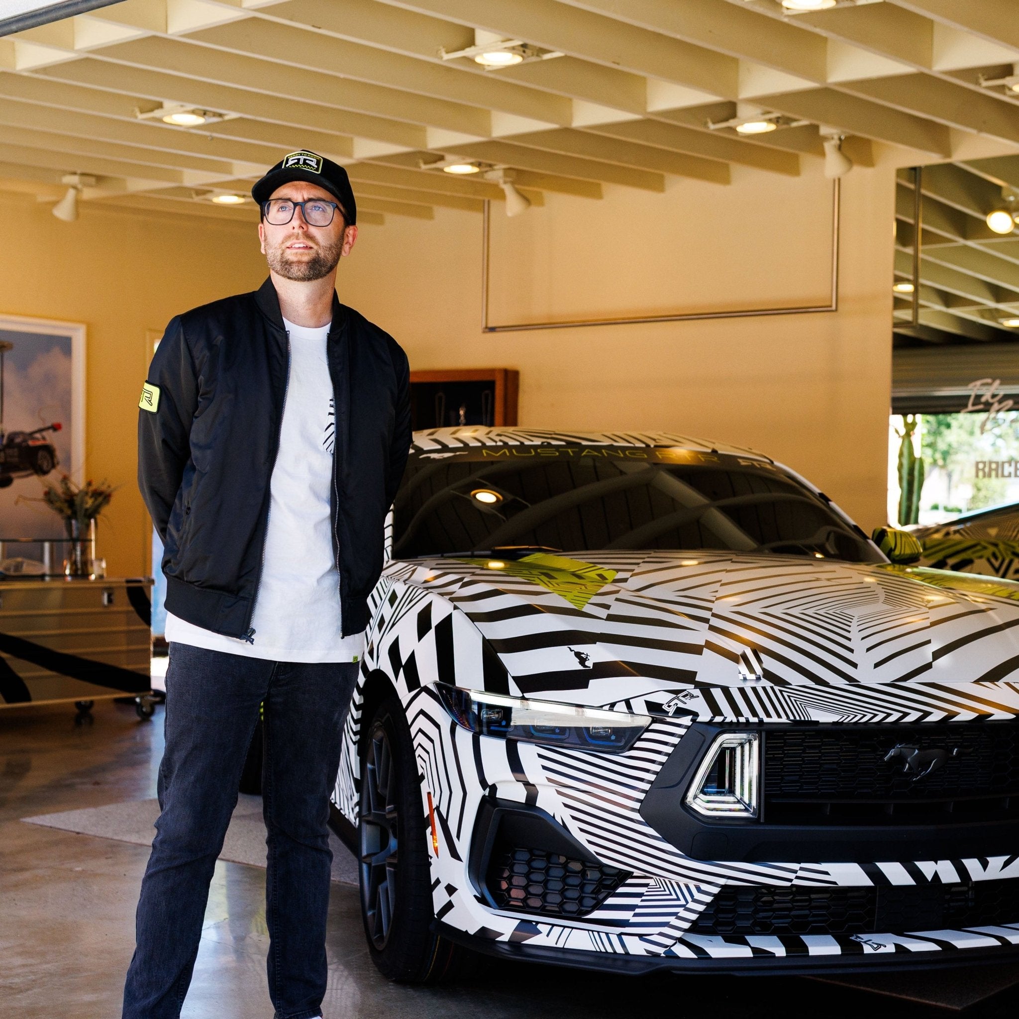 Man in Ford Mustang x RTR Collection Stealth Jacket stands by a geometric-patterned Mustang, highlighting the jacket's performance-inspired design and modern utility.