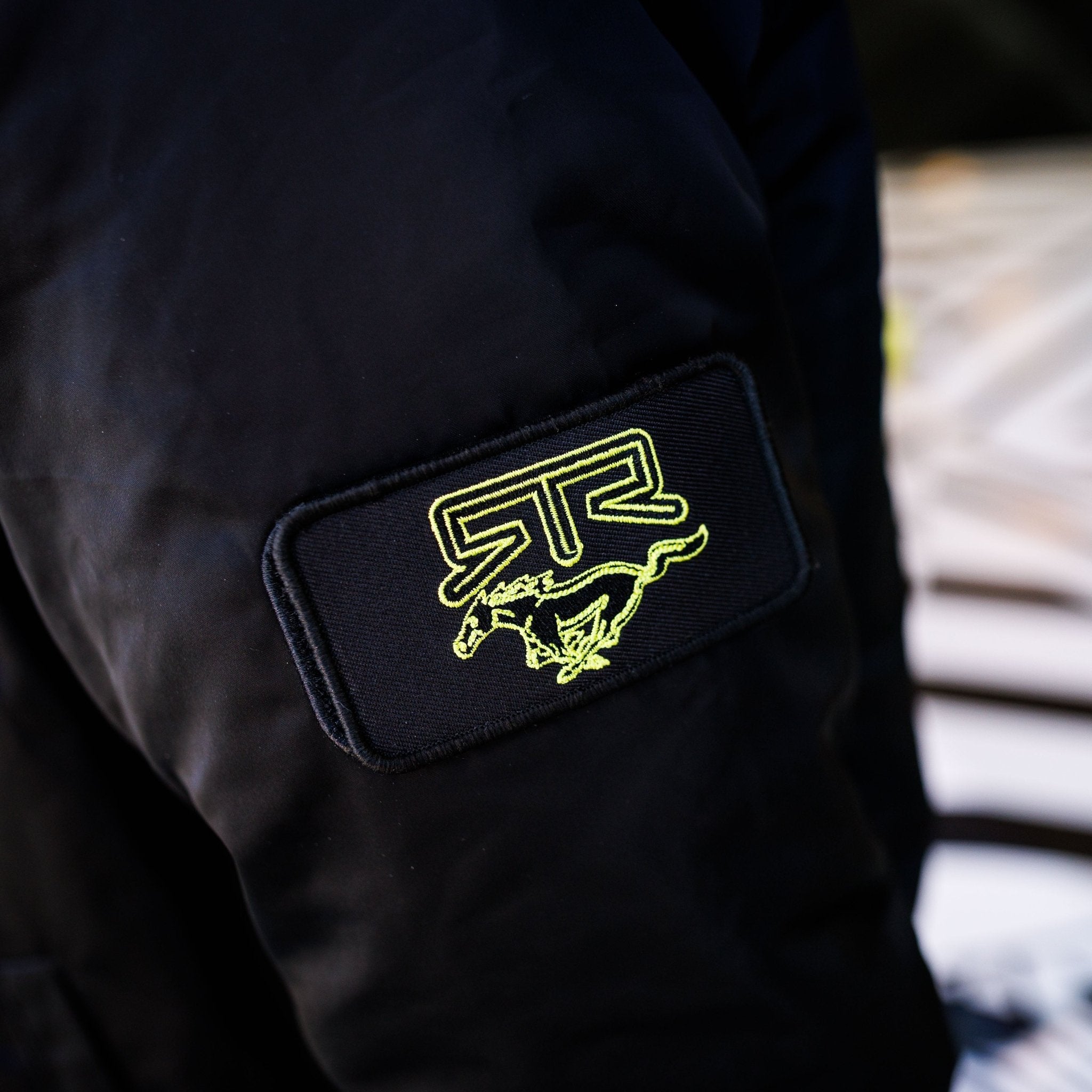 Close-up of the Ford Mustang x RTR Collection Stealth Jacket sleeve with “RTR” and a horse outline on a black patch in yellow-green embroidery.