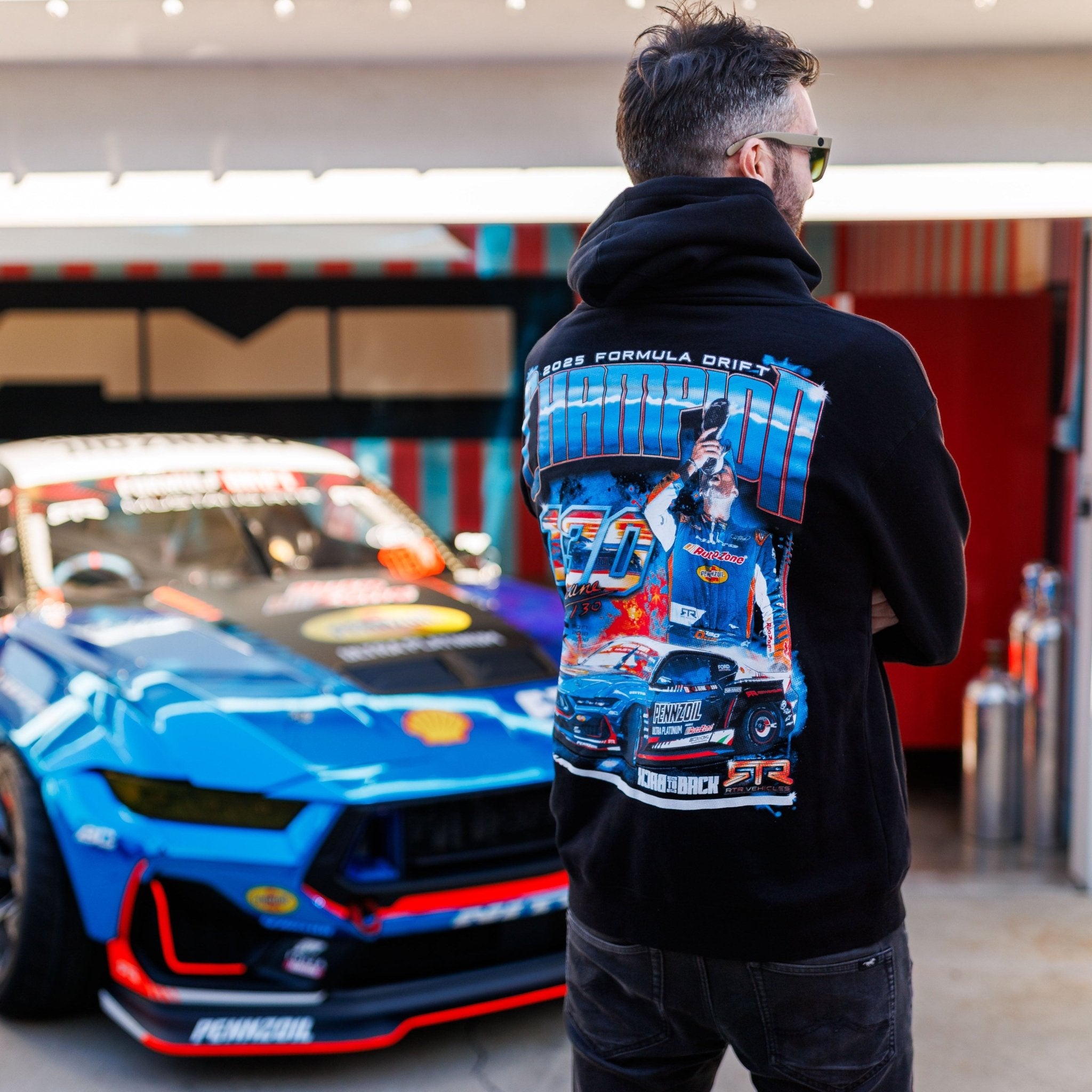 Man wearing James Deane 2025 Championship Hoodie in race-car paddock, featuring vibrant drifting graphics, stands near a modified blue drift car with sponsor logos.