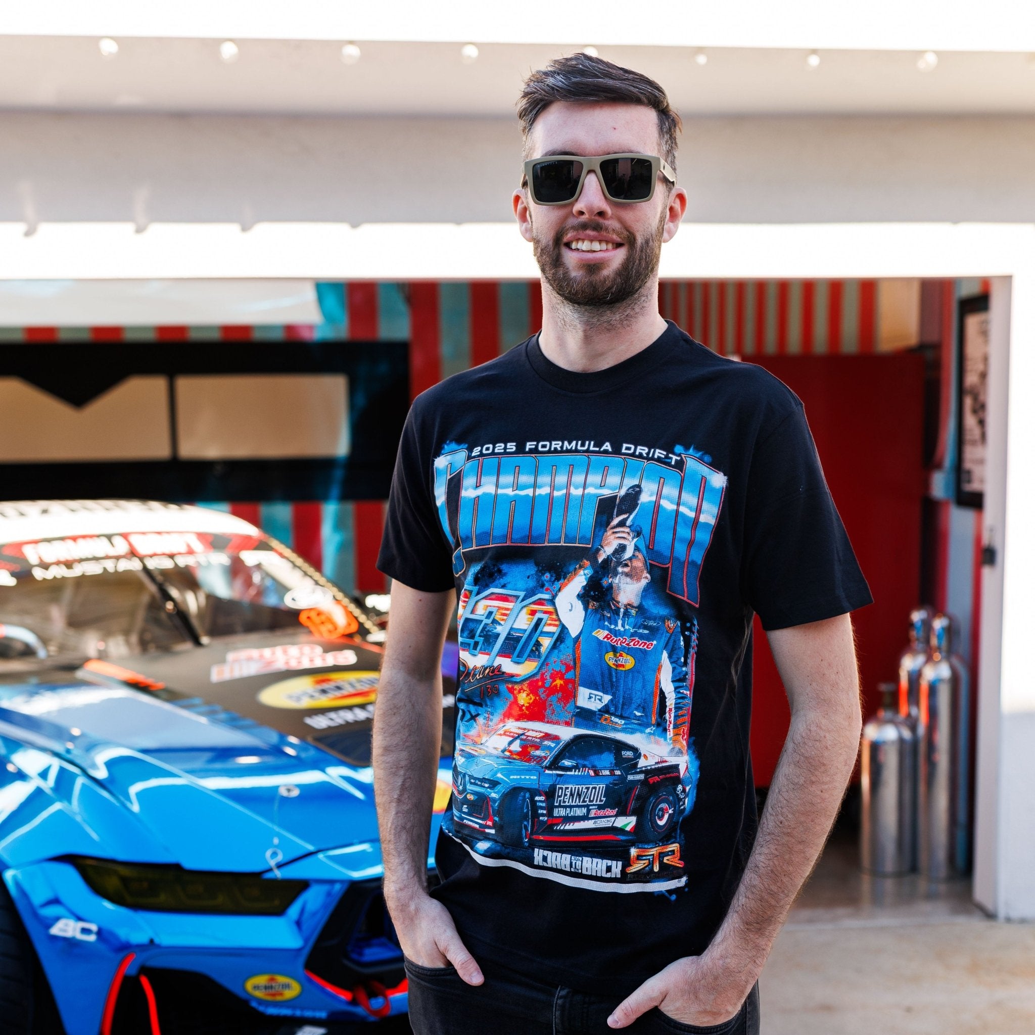 James Deane 2025 Championship T-Shirt featuring champion graphics and sponsor logos, worn by a young man beside a blue drift car in a garage.