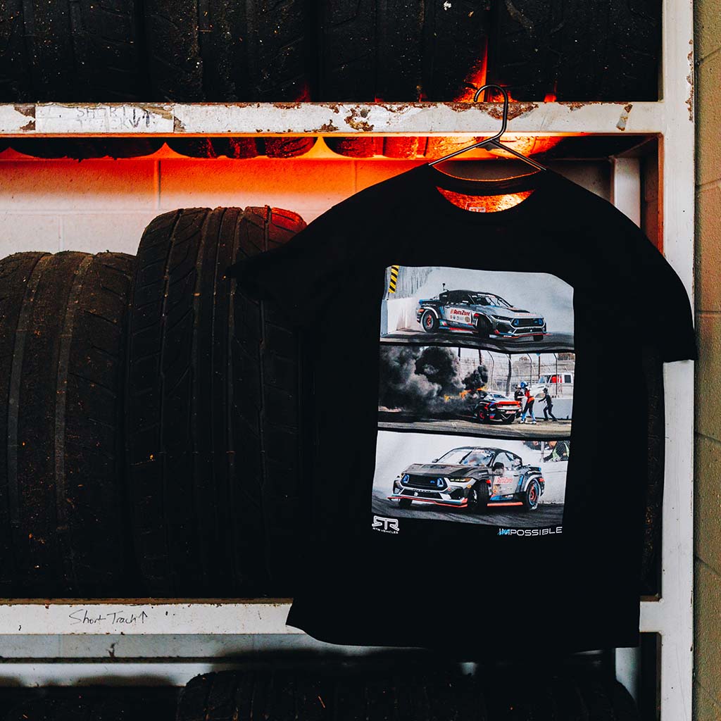 Never Quit Tee on a tire rack featuring James Deane's car photos, smoke engulfed track image, and handwritten Never Quit message. Limited-edition Formula Drift memorabilia for RTR fans.