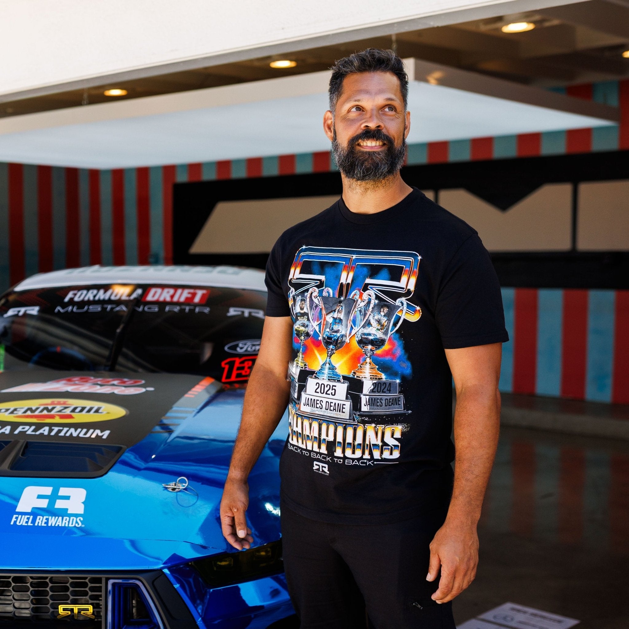 A bearded man wearing the RTR Back To Back Champions T-Shirt stands beside a branded Mustang, celebrating RTR's Formula Drift victories.