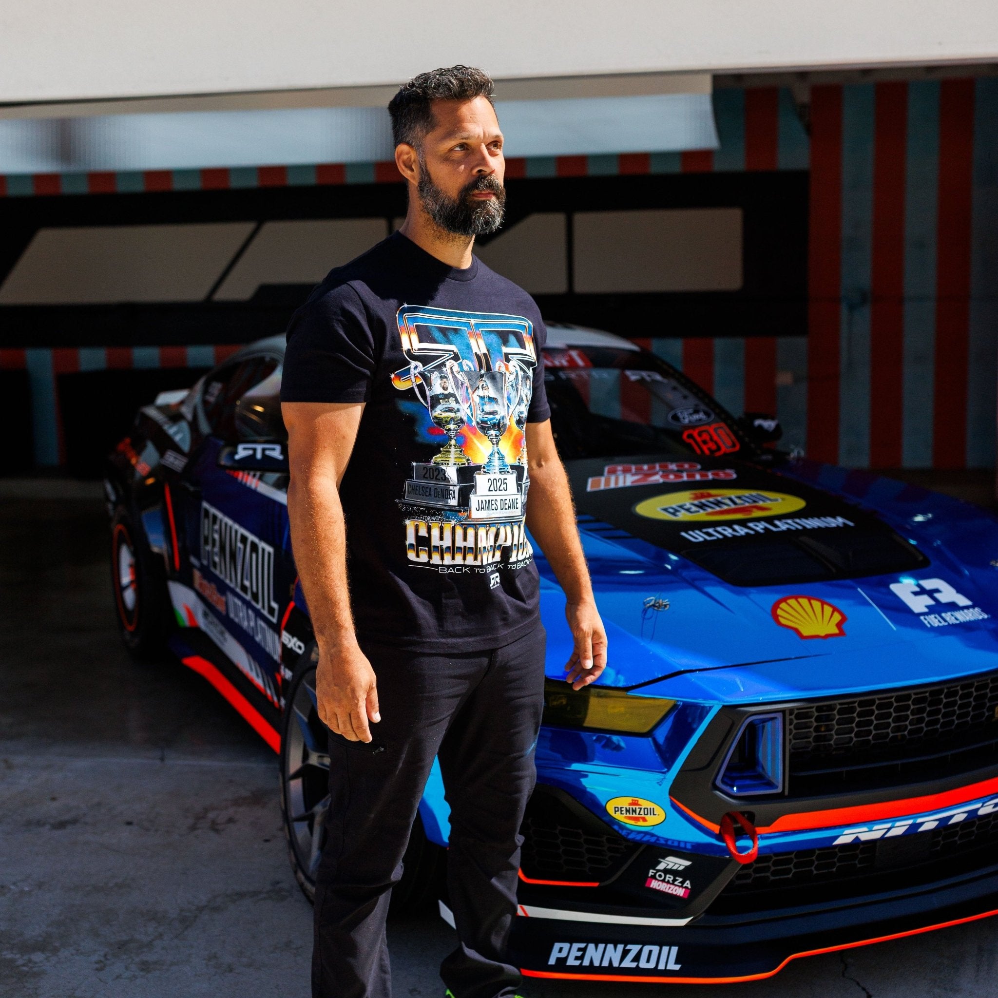 Man in RTR Back To Back Champions T-Shirt stands by a Pennzoil-Shell race car, celebrating RTR's Formula Drift dominance with trophy graphics and championship details.