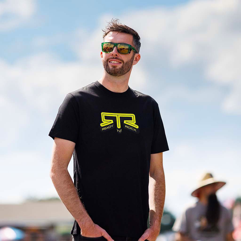 James Deane wearing a black RTR Core T-Shirt with yellow RTR logo outdoors.