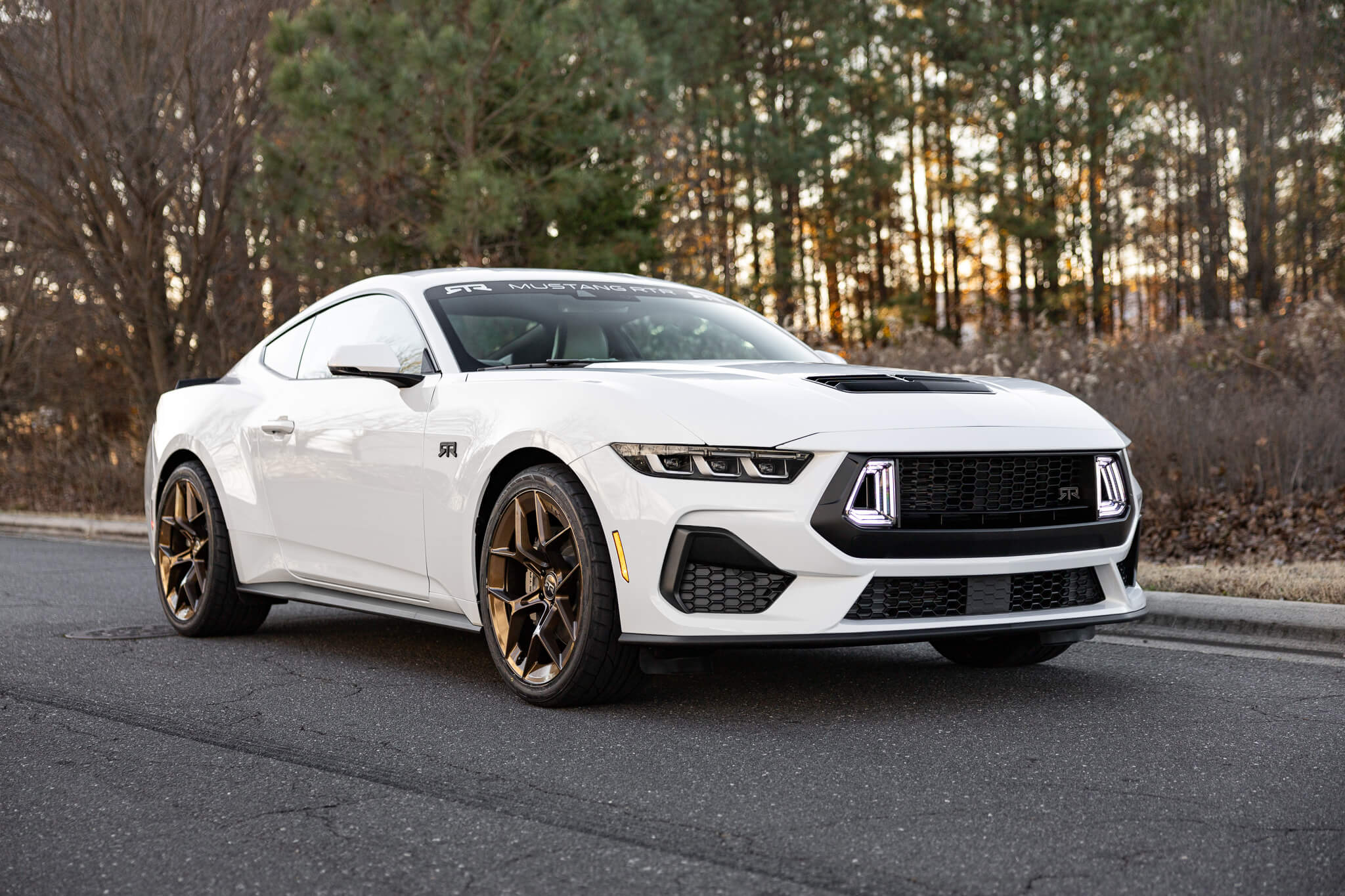 A white 2024 Ford Mustang GT with RTR upper grilles and LED air intakes, parked on a street with trees in the background.