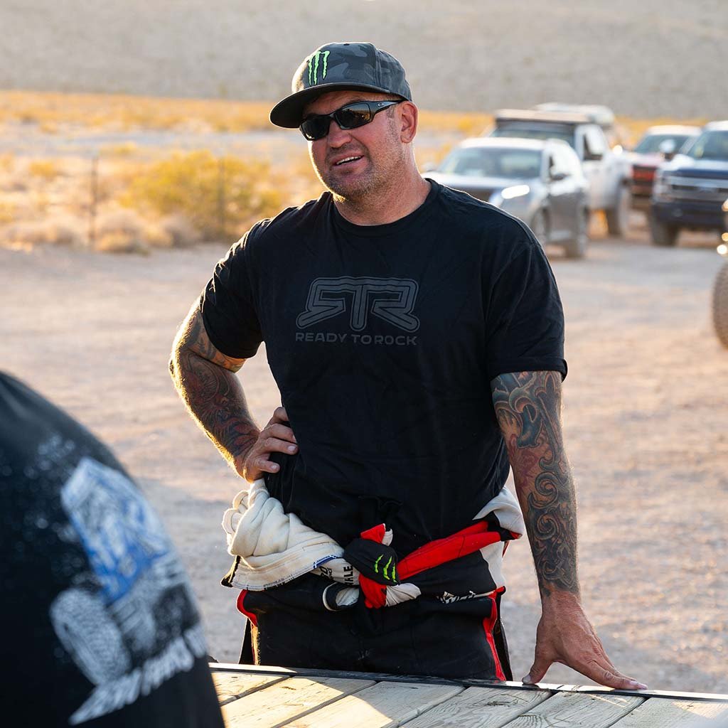 Man wearing a black RTR Tonal t-shirt in an outdoor setting with vehicles in the background