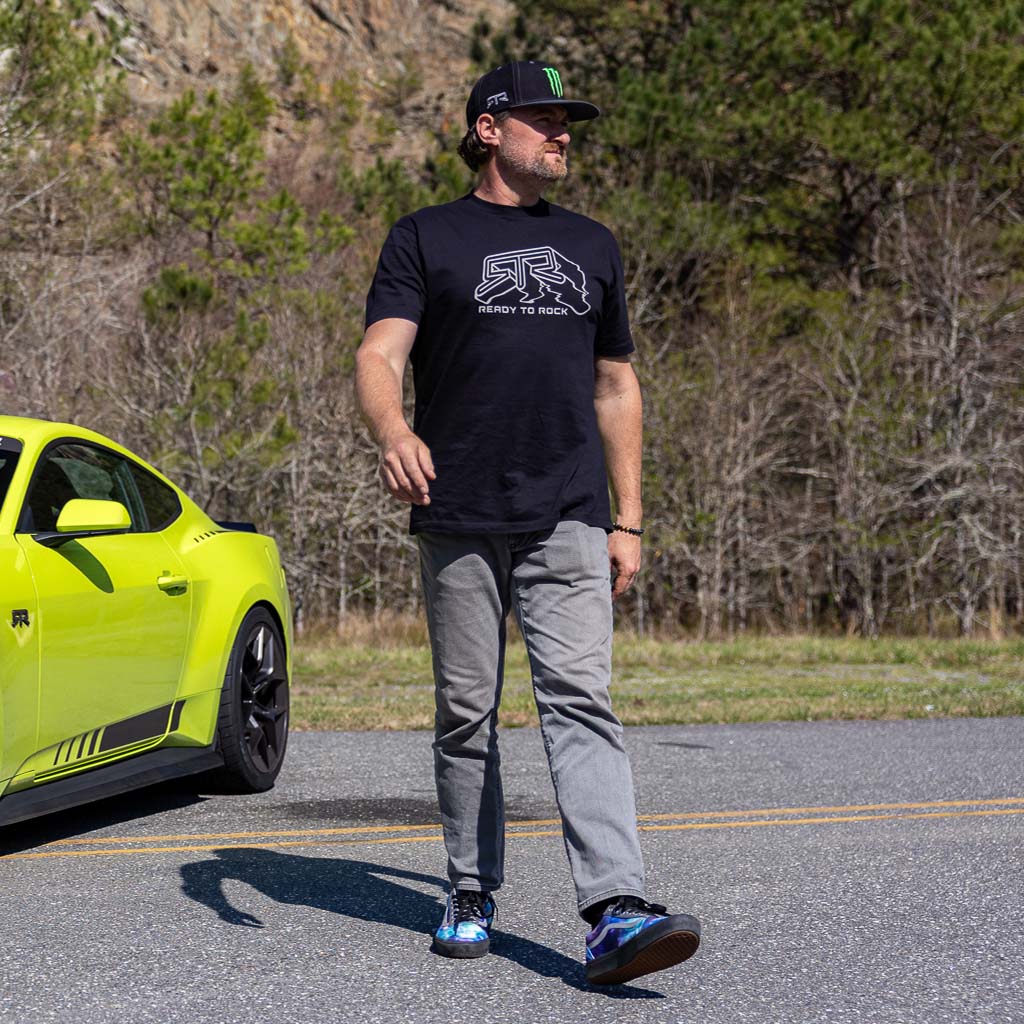 A man stands next to a car, wearing the RTR Warped T-Shirt featuring a 3D distorted RTR logo design, highlighting its dynamic track-inspired style.