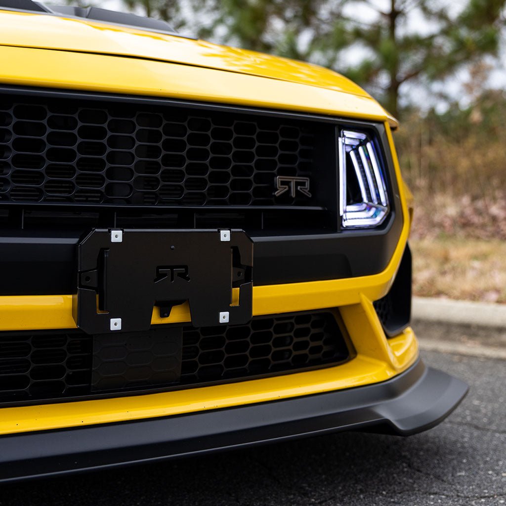 Close-up of RTR Front License Plate Bracket on yellow Mustang with RTR Upper Grille, showcasing sleek black aluminum finish and RTR logo.