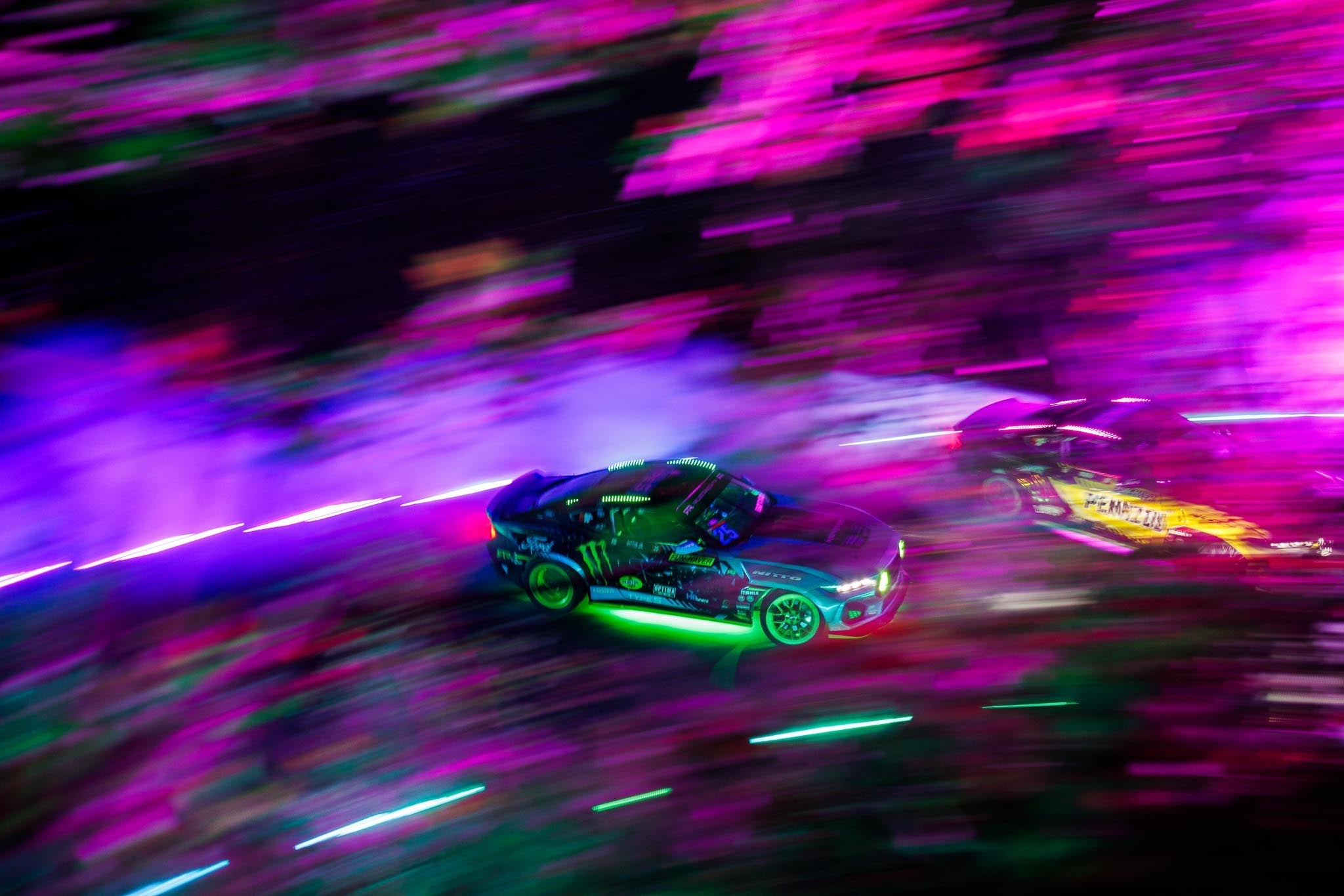 Two vibrant Mustang RTR Spec 5-Ds drifting on a mountain road, featuring lighting with green, magenta, and violet hues.