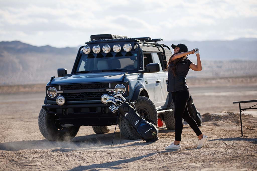 A woman swings a golf club near a Bronco RTR ROVR on a dirt road, with a bag of golf clubs nearby.