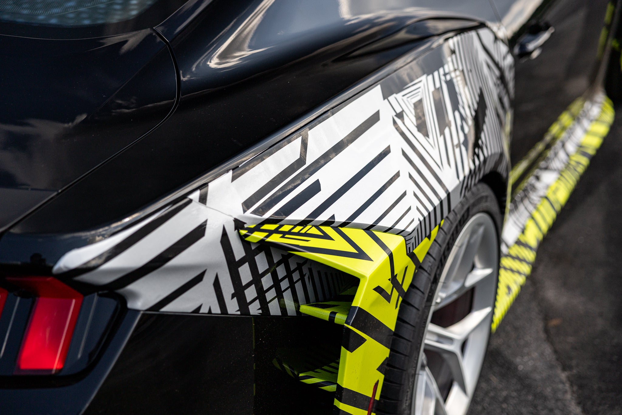 Close-up of a black 2026 Mustang RTR Spec 5 with a hyperlime and black camouflaged rear quarter panels on a road.