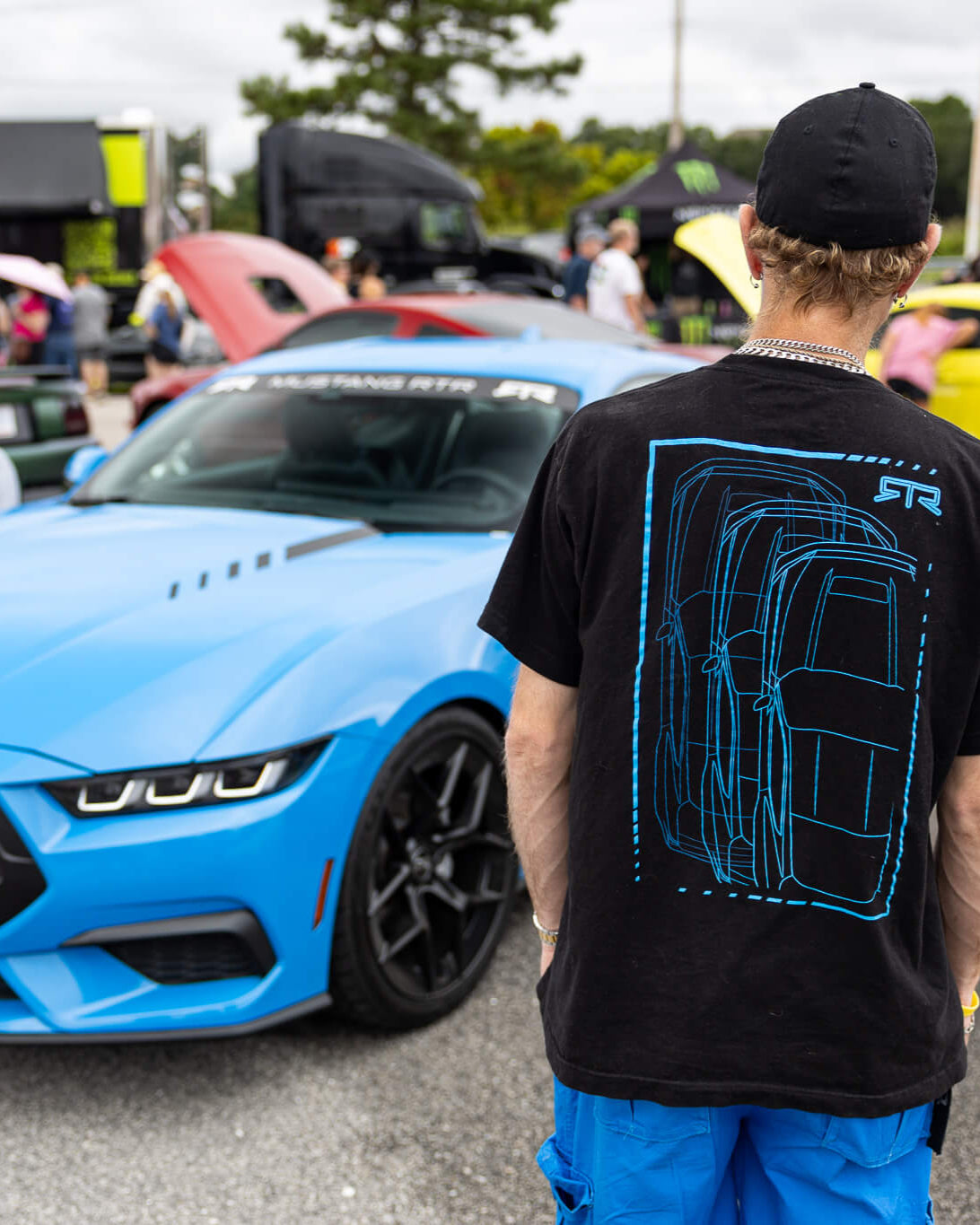 Person wearing a black t-shirt with a car design at a car show with a grabber blue Mustang RTR in the background.
