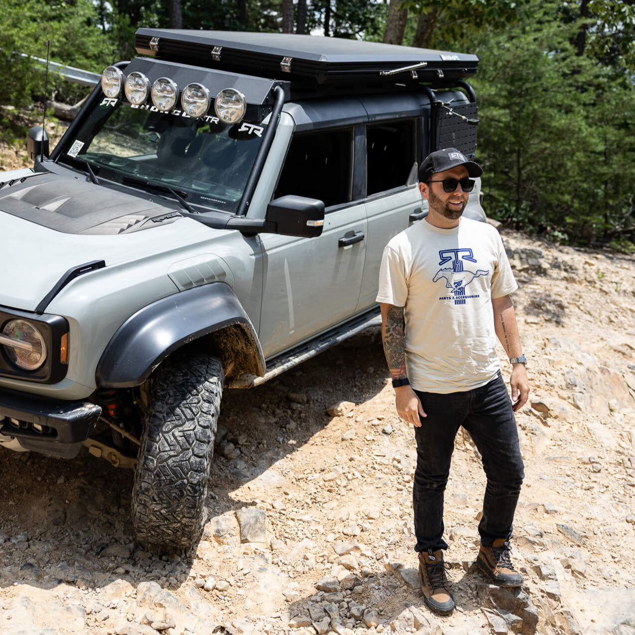 Man standing next to a gray off-road Bronco on a dirt trail with trees in the background