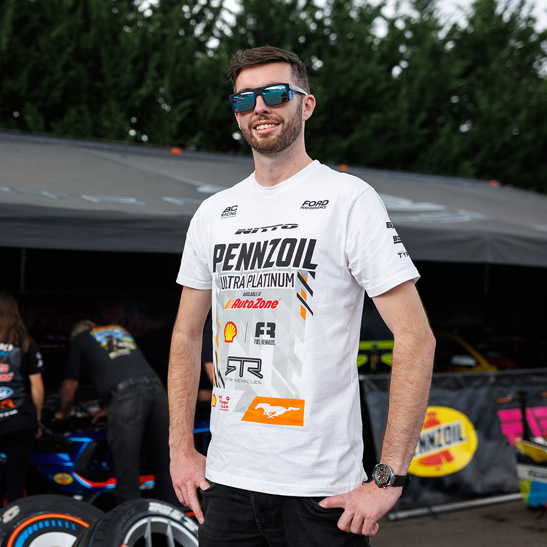 James Deane in his Pennzoil team shirt standing in front of a racing car garage.