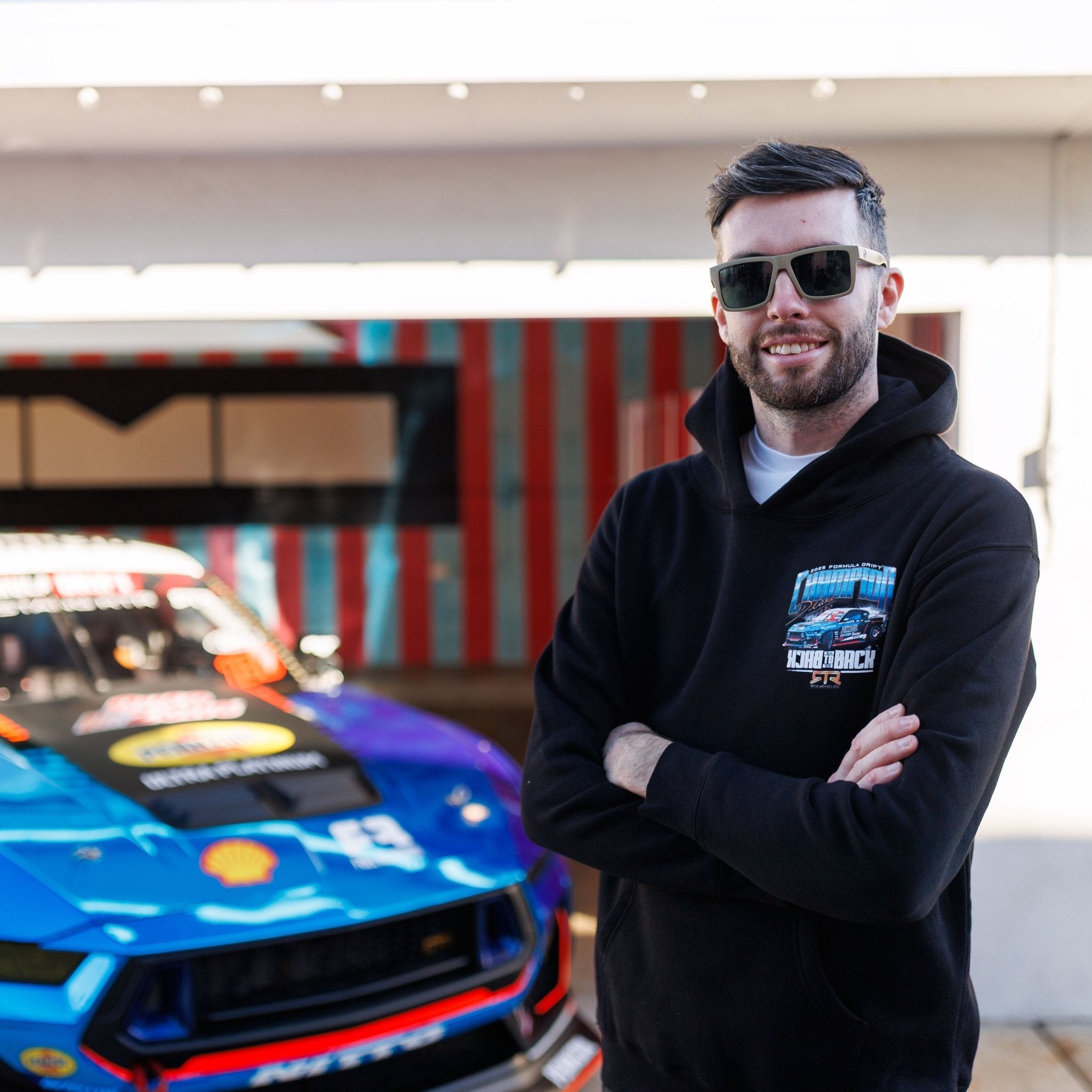 Man in sunglasses wearing James Deane 2025 Championship Hoodie, standing confidently with arms crossed in front of a sponsor-covered race car.