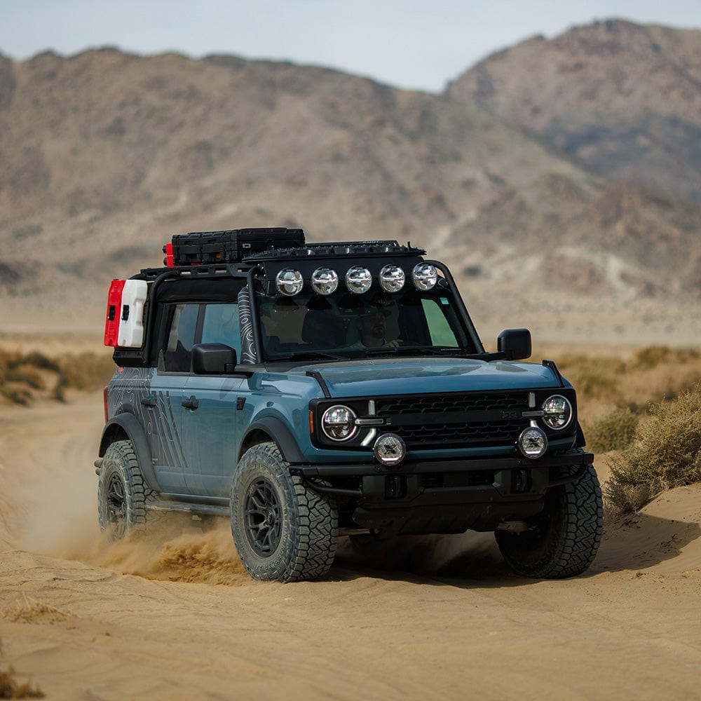 RTR Roof Rack & RTR Light Bar with PROJECT X Lights for 21+ Bronco, 22+ Bronco Raptor on a blue Bronco driving off-road. Sturdy steel construction with integrated light bar for adventure.